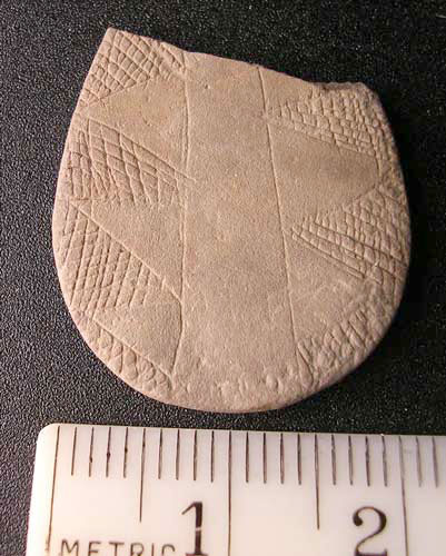 stone-like artifact with a chip missing measuring 2" across, indented in triangle-shapes and cross-hatched