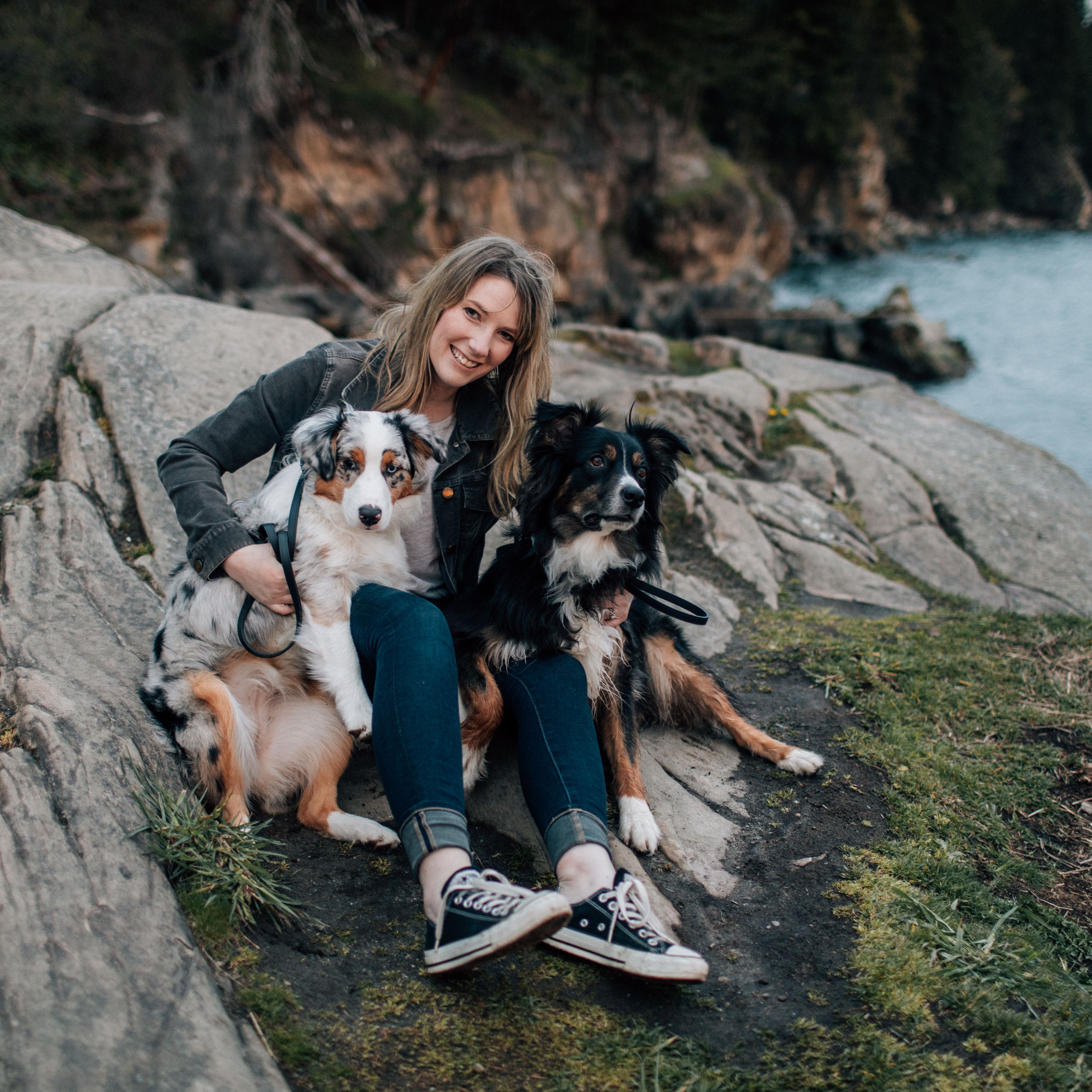 courtney sitting and hugging her two fluffy dogs on the coastline with the ocean in the background