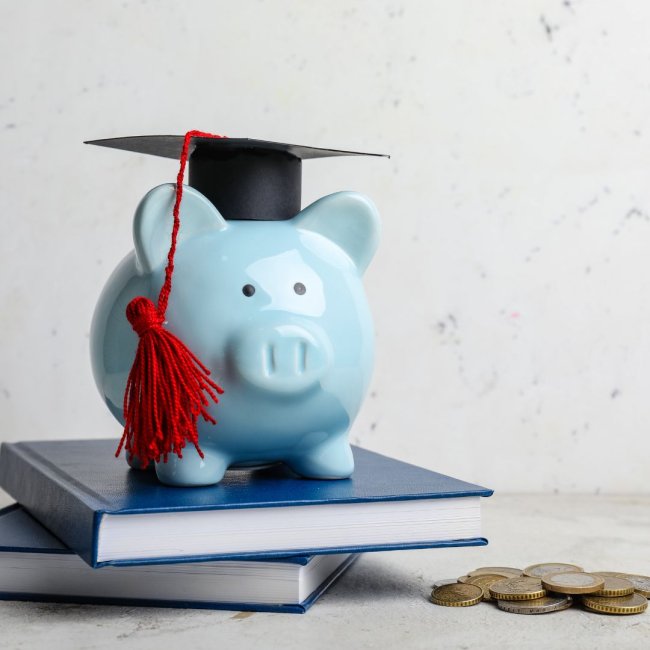 Blue piggy bank with graduation cap on top of two books with coins spilled out next to it.