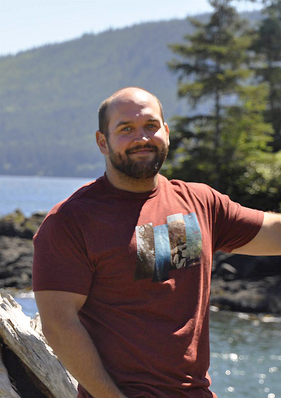 Steve Bennett wearing a reddish orange t-shirt with a graphic on it standing near a glistening mountain lake