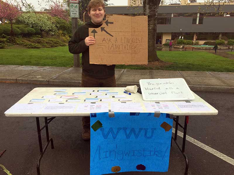 a student tabling while holding a cardboard sign that says "ask a linguist anything"