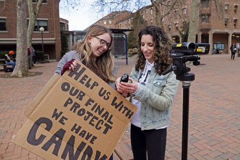 Two students holding a cardboard sign reading "Help us with our final project, we have candy"