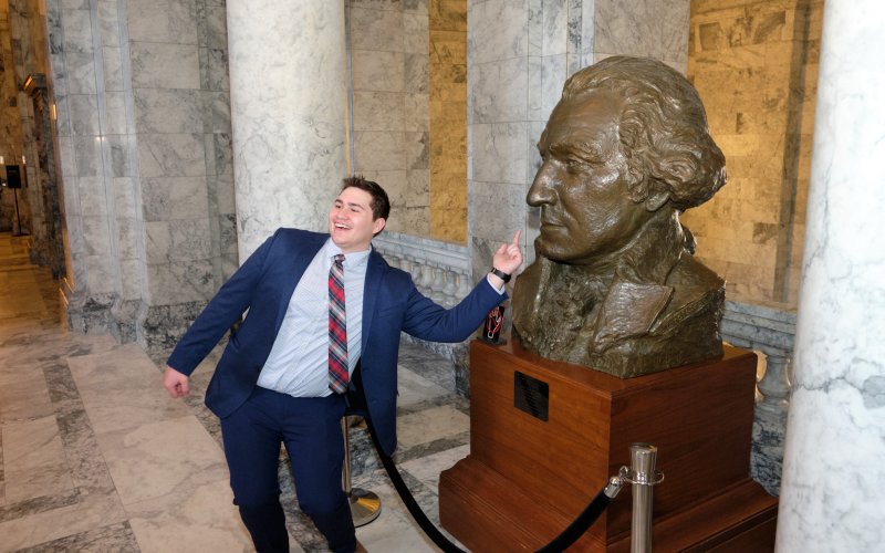 Student posing in excitintment while pointing towards a bust of George Washington