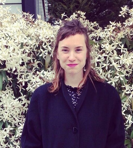 Ruthie wearing long winter coat and standing outside in front of flowering plant