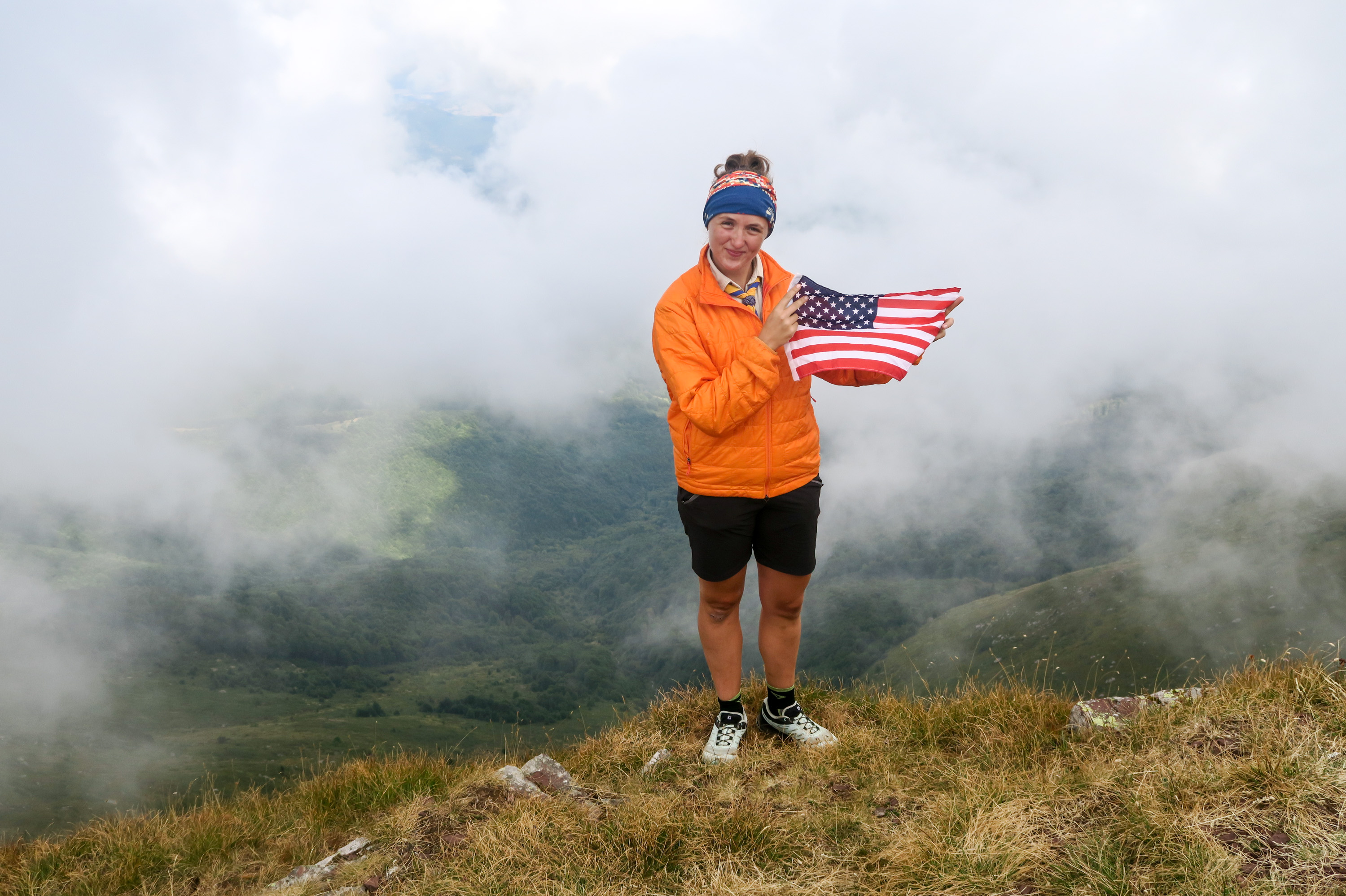 Carla standing atop mountain, holding USA flag, cloudy skies in background