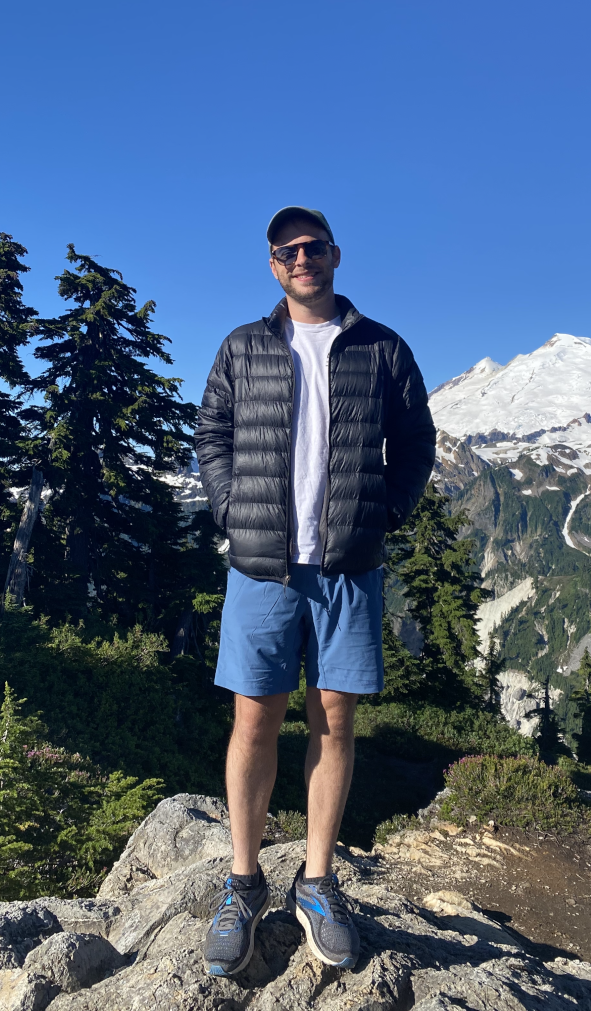 Patrick standing on rock with his hands in his pocket and smiling at the camera, mountains and trees behind him