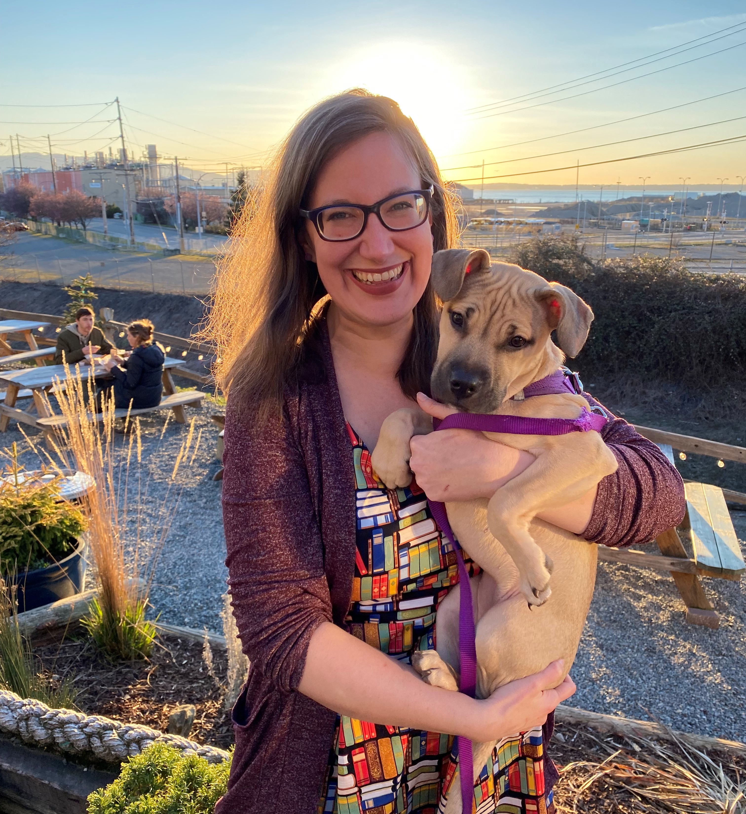 Smiling woman standing in front of sunset with brown dog in her arms