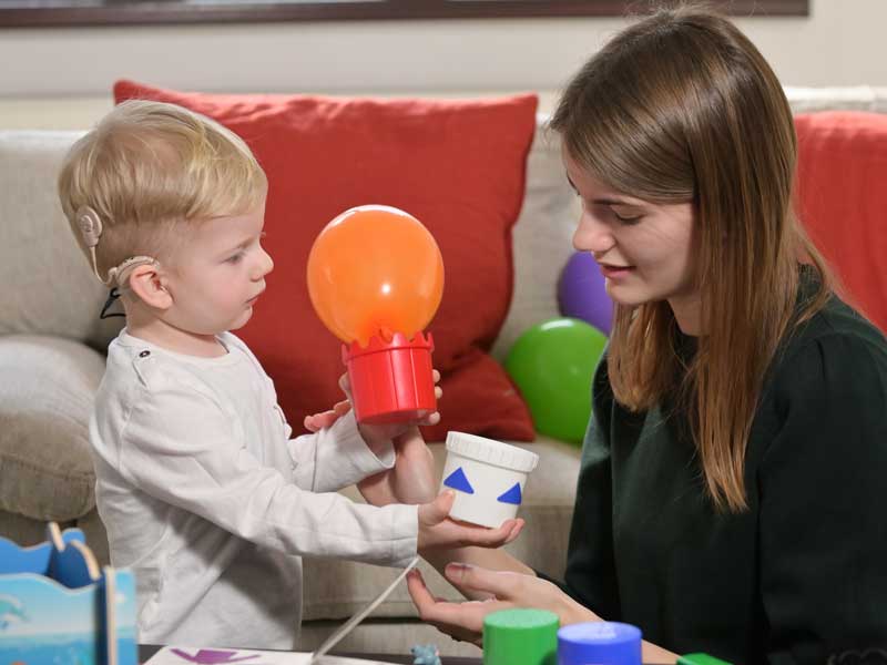 A child wearing hearing aids plays with brightly colored toys near a colorfully decorated sofa. A woman sits beside the child engaging with them.