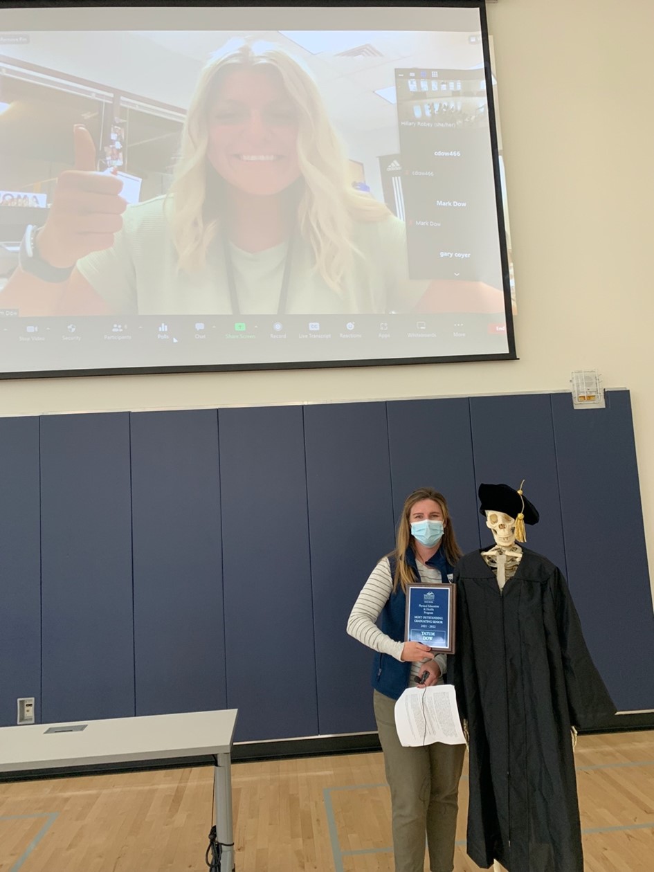 Professor, model skeleton in graduation robes smiling and standing in front of projector screen picture of smiling student