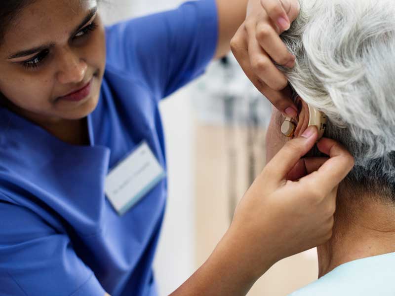 A medical professional in blue scrubs fits an older patient with hearing aids in a clinical setting.