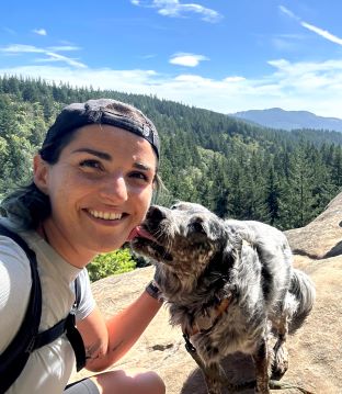 Marianne on a hike/mountain peak being licked by her dog. 