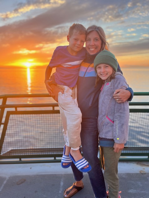 Lindsay and two kids standing on ferry deck in front of a sunset 