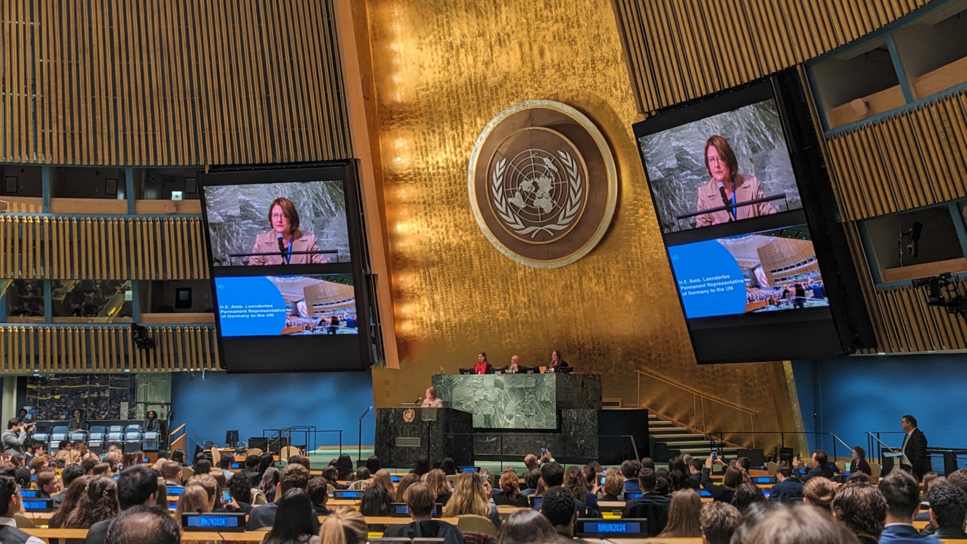  In the United Nations General Assembly Hall, a woman stands at a podium, speaking into a microphone. She is wearing a suit and has short brown hair. There is an emblem on the wall behind her. The hall is filled with people, mostly young people, who are sitting in rows of chairs. They are listening to the woman speak. On either side of the woman there are two large screens which show a close up of her face.