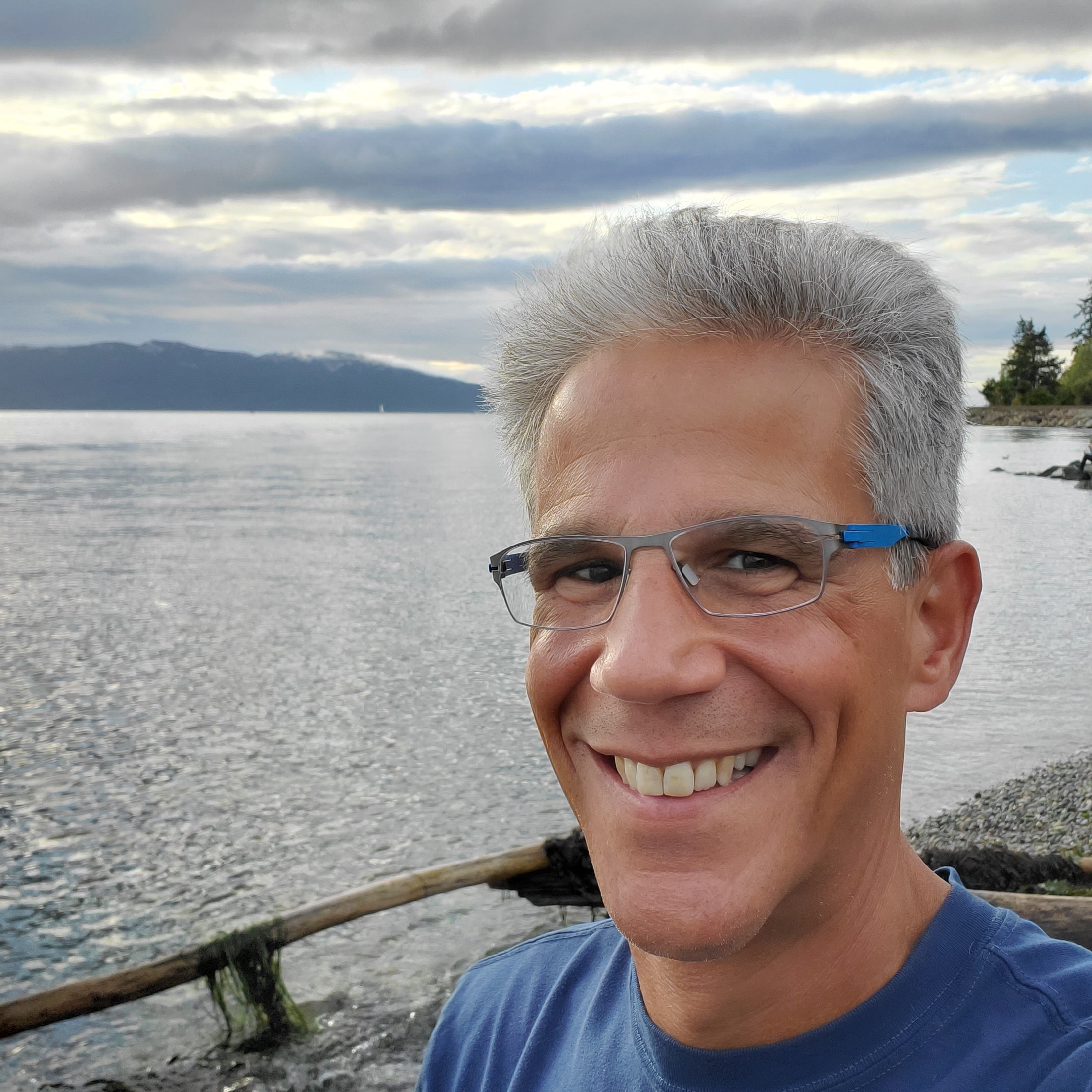 Headshot of Marc Geisler smiling wearing with a blue shirt and glasses with a waterfront in the background.