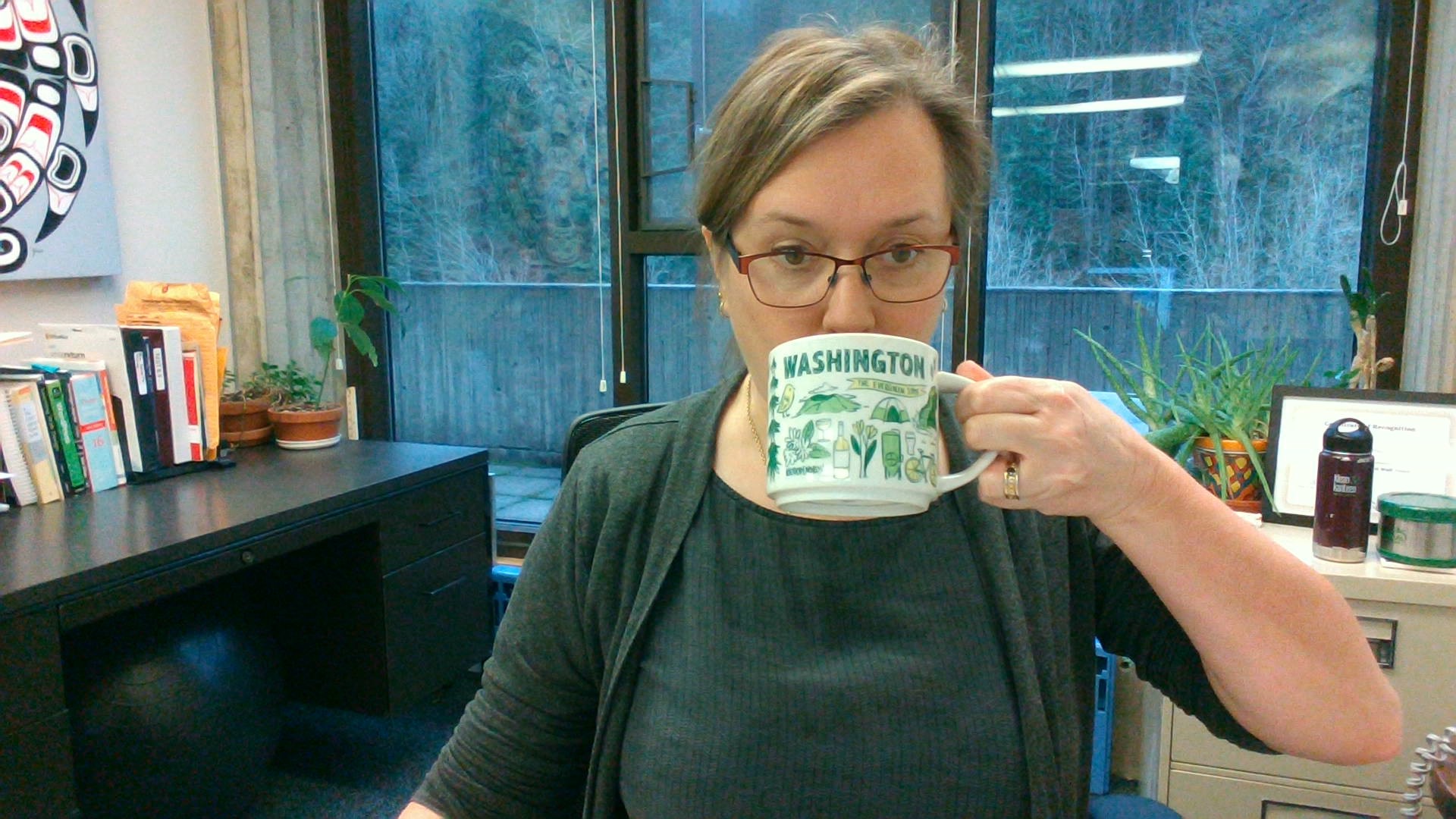 Lauren Townshend drinks from a large coffee mug in the office, which overlooks a forested area. 