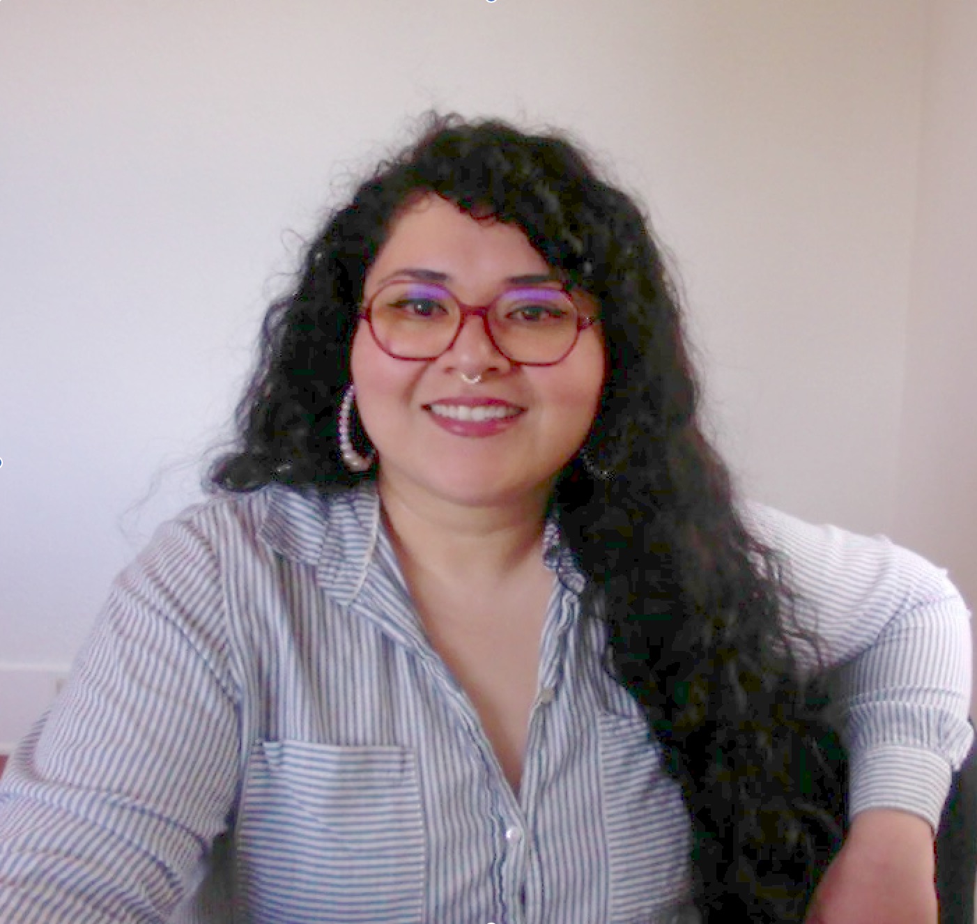 Melina smiling and looking at the camera. She is wearing a white blue-stripped blouse, long brown hair, and glasses.