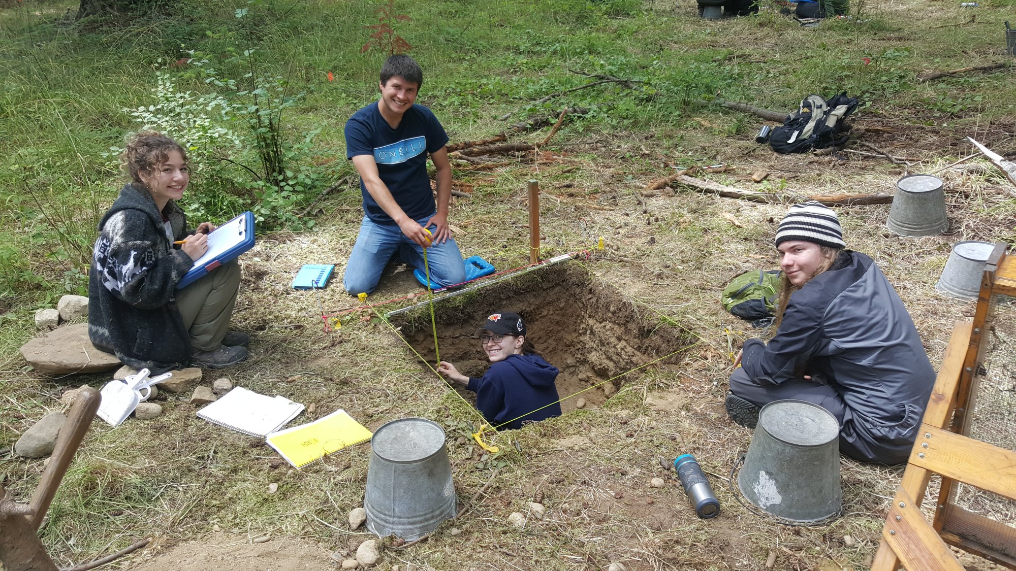 students participating in a dig, sitting near a square hole surrounded by tools