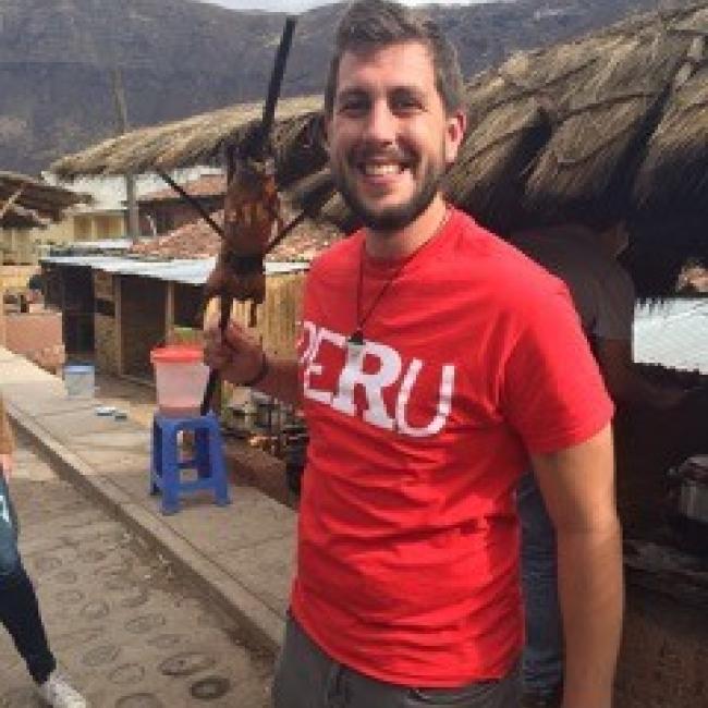 Jacob wearing red tshirt that reads Peru, holding cuy (guinea pig) on a stick, mountains in background, located in Peru. 