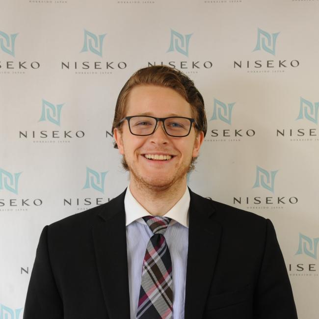 Mitchel wearing suit and tie, standing in front of wall that has  the words Niseko displayed