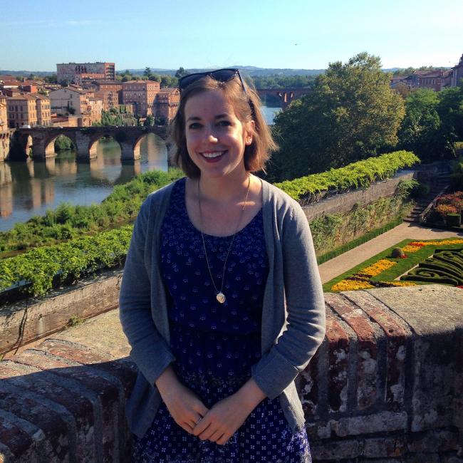 Anneliese standing outside along a brick walkway on a sunny day, with river and buildings in background. Location is Albi, France.