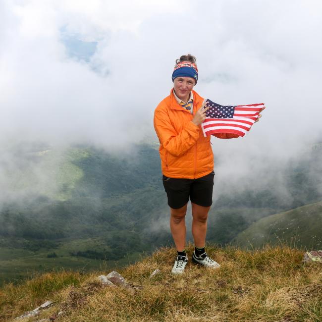 Carla standing atop mountain, holding USA flag, cloudy skies in background