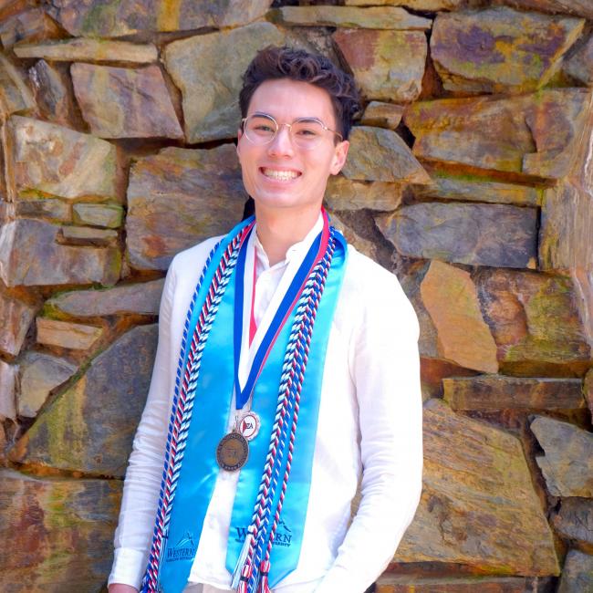 Nate Jo wearing a sash, braided tassels, and a medal in front of a stone wall