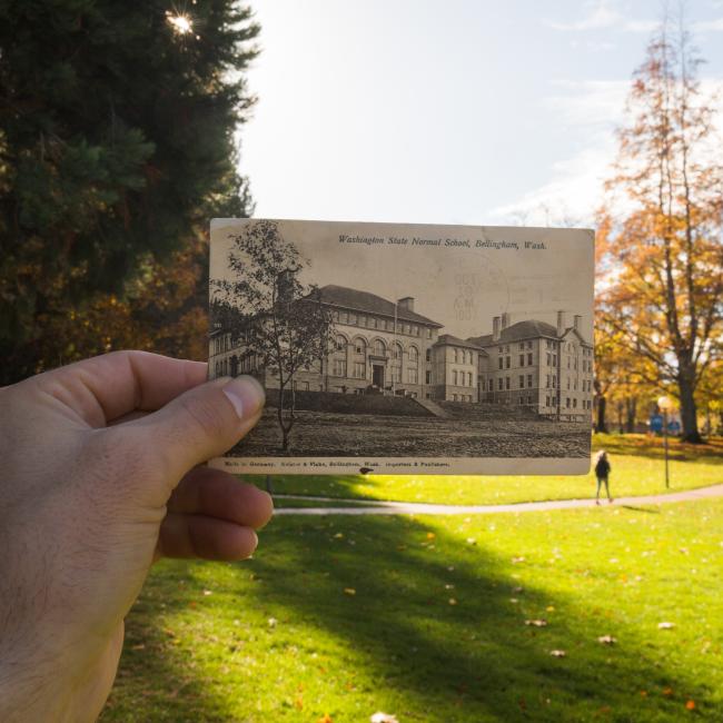 hand holding a postcard image of Old Main in front of the present standing Old Main