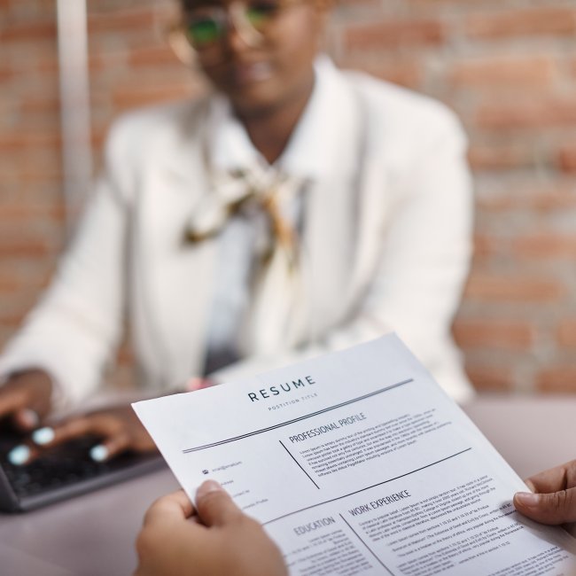 A person holding a resume and in the background we see another person who is out of focus using a laptop.