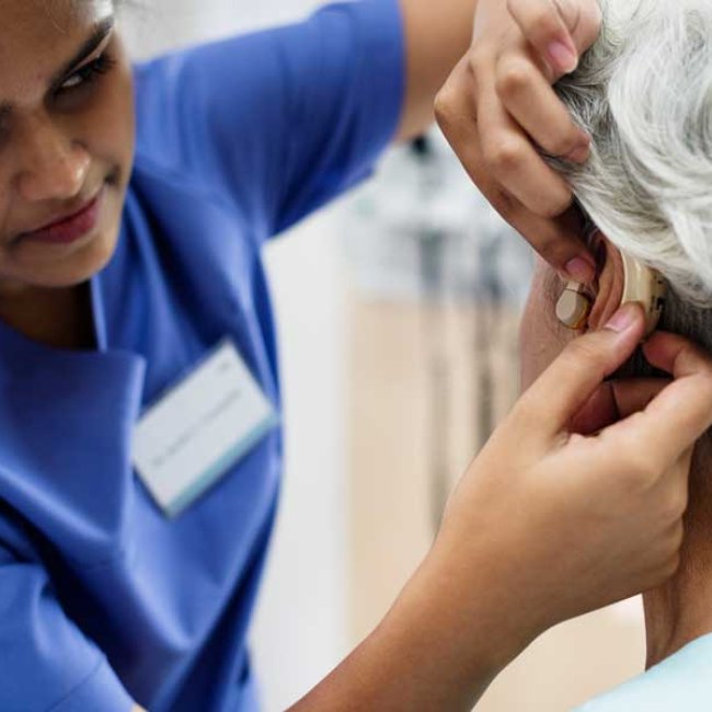 A medical professional in blue scrubs fits an older patient with hearing aids in a clinical setting.