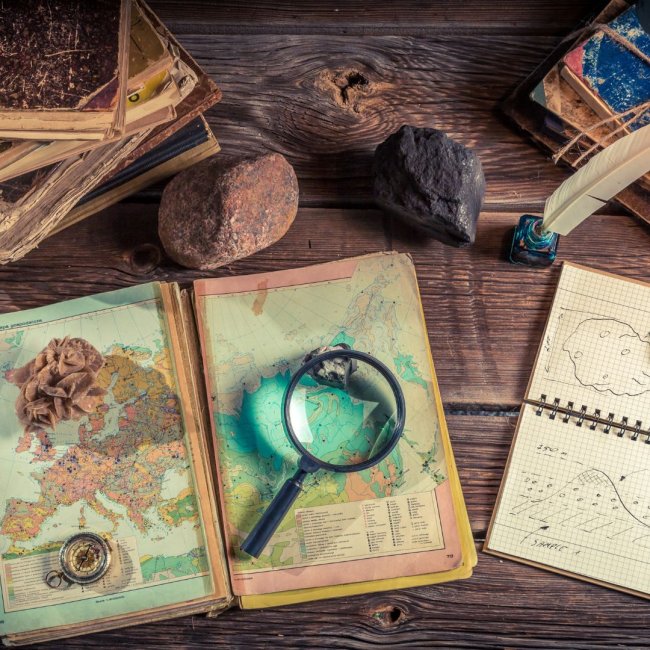 Old books, maps, rock, compass, magnify glass, and notes on a desk