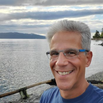 Headshot of Marc Geisler smiling wearing with a blue shirt and glasses with a waterfront in the background.