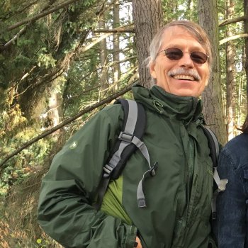 James Loucky wears a green jacket, gray backpack, and sunglasses while smiling in the woods