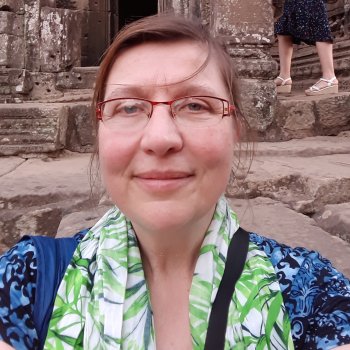 Judith takes a selfie in front of a stone building. She wears a patterned blue shirt, a scarf with a leaf pattern, and glasses.
