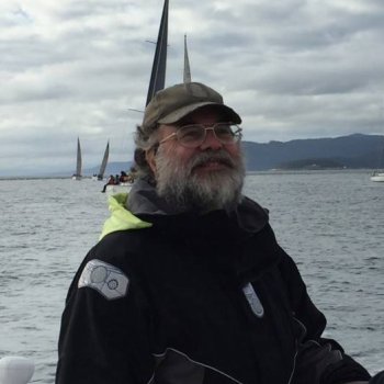 Todd wears a all-weather jacket, gray cap, and a gray beard. Behind Todd is a body of water with boats and land masses in the distance