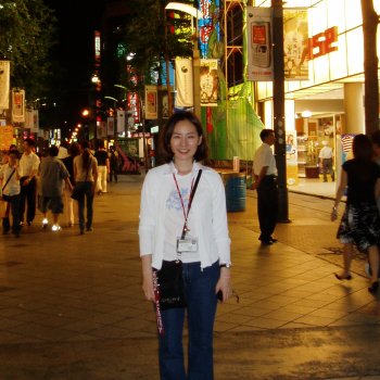 Yeon Jung stands on a busy street at night, wearing a conference badge and a white sweater. The building behind Yeon is lit brightly.