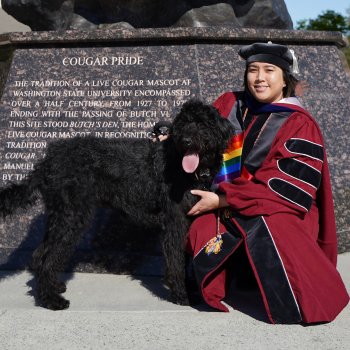 Dr. Paul/Leena/Paulina Abustan (any pronoun) gender fluid Pilipinx person with light brown skin wearing WSU PhD red and black graduation regalia with a rainbow stole with their black access labradoodle, Meelo
