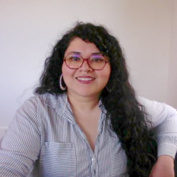 Melina smiling and looking at the camera. She is wearing a white blue-stripped blouse, long brown hair, and glasses.