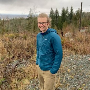 Zander Albertson, who has light skin and long blond hair tied back, wearing glasses and a teal windbreaker hanging out in nature