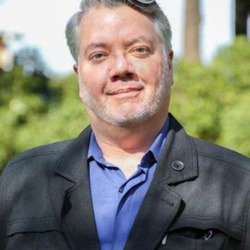 Troy Abel, who has is light skinned with short gray hair and short facial hair, wearing a black sport coat over a blue shirt on a sunny day 