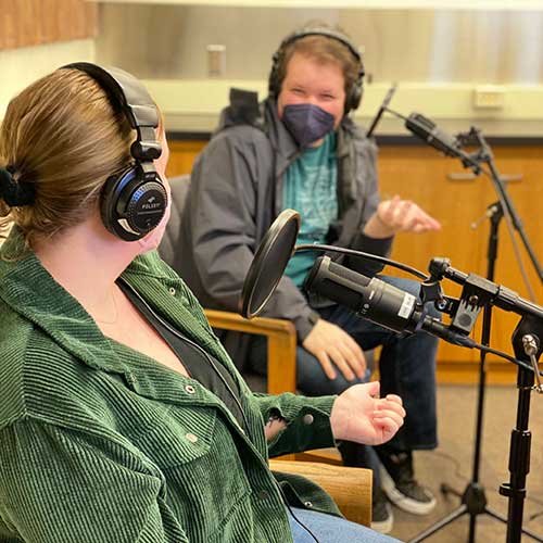 From left, WWU students Emma Ahmann and Niko Attebery recording The Podling podcast. Both are wearing headphones and talking into microphones.