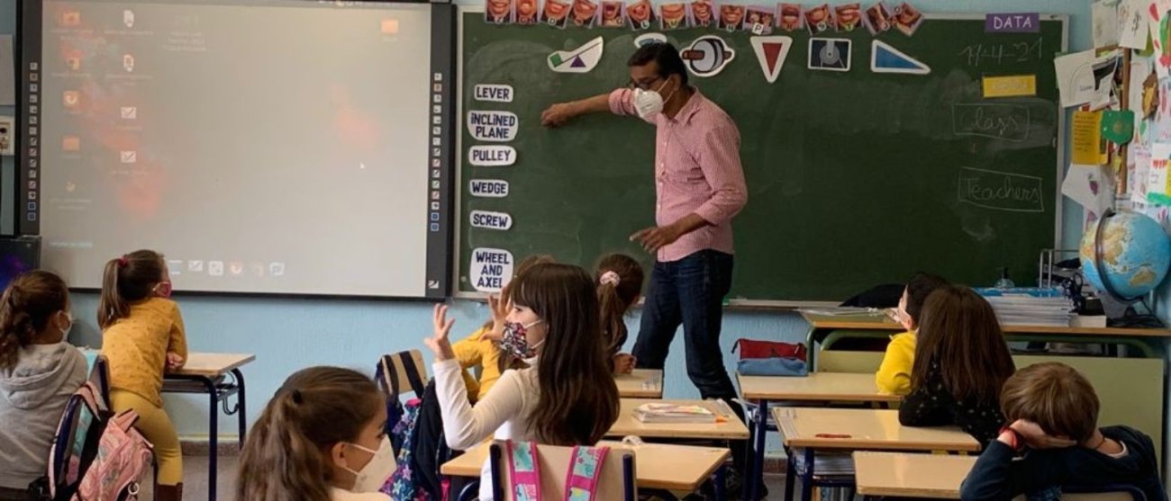 A teacher wearing a pink shirt and a mask standing in front of a class of young kids. The teacher is pointing at the chalkboard while interacting with the kids in the class.