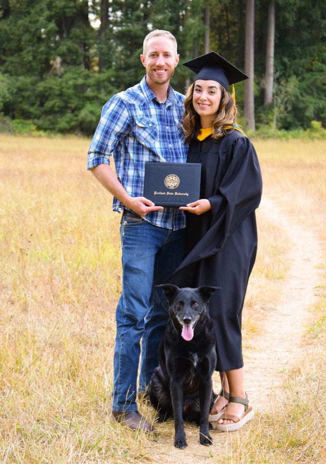 Smiling man and smiling woman in graduation robes holding a degree standing in a field with a dog sitting at their feet