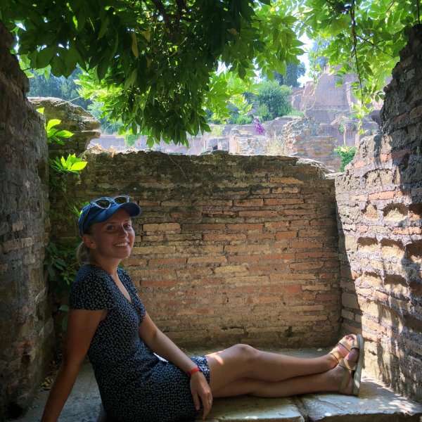 Skyla Sorensen in a polka dot dress and blue baseball cap sits in a tree-shaded stone nook in Italy
