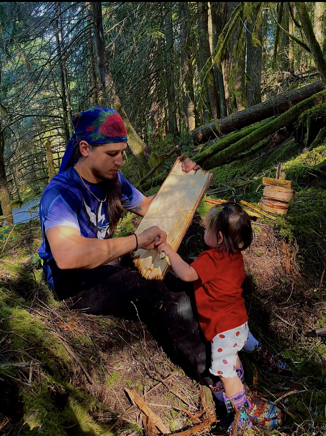 A young person wearing a multicolored bandana teaching his less than one-year-old daughter how to clean cedar bark and make stuff.