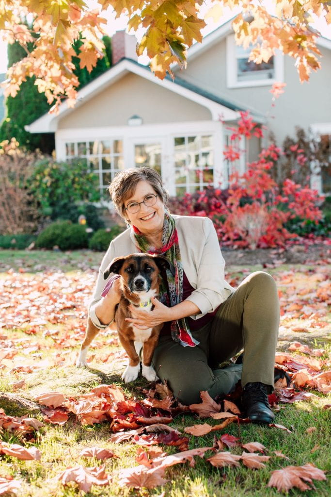 Brenda Miller holding her dog, Barnaby, on a lawn covered in red and orange fallen leaves.
