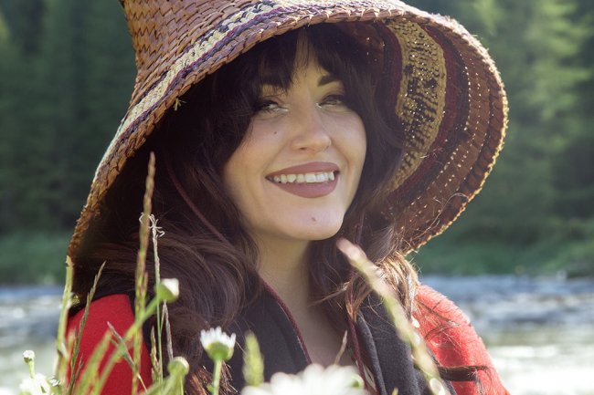  A young woman wearing a traditional Chilkat hat and red shawl smiles in a field of daisies.